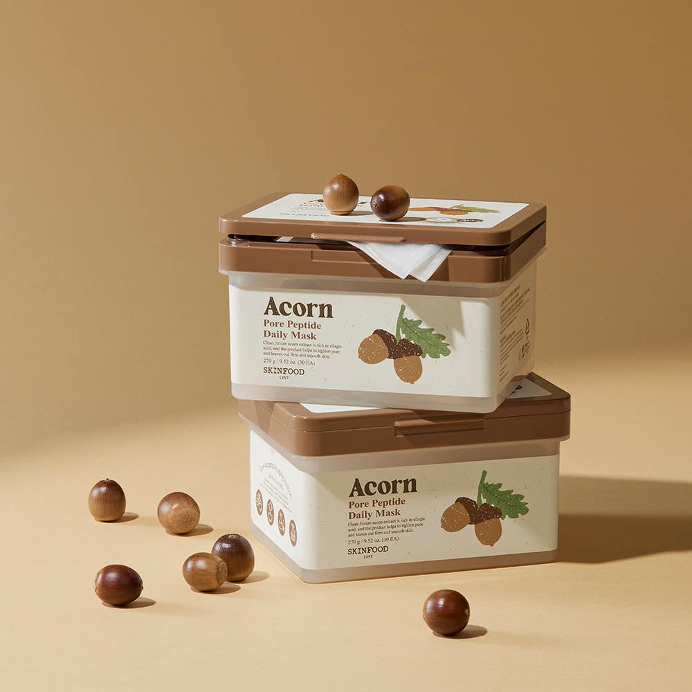 Two beige and brown skincare containers with acorn illustrations stacked on a tan surface with scattered acorns nearby