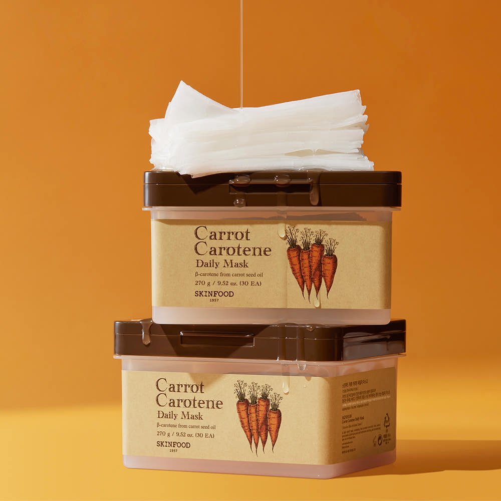 Two beige skincare containers with brown lids stacked, labeled with carrot illustrations and white mask sheets on top against orange backdrop