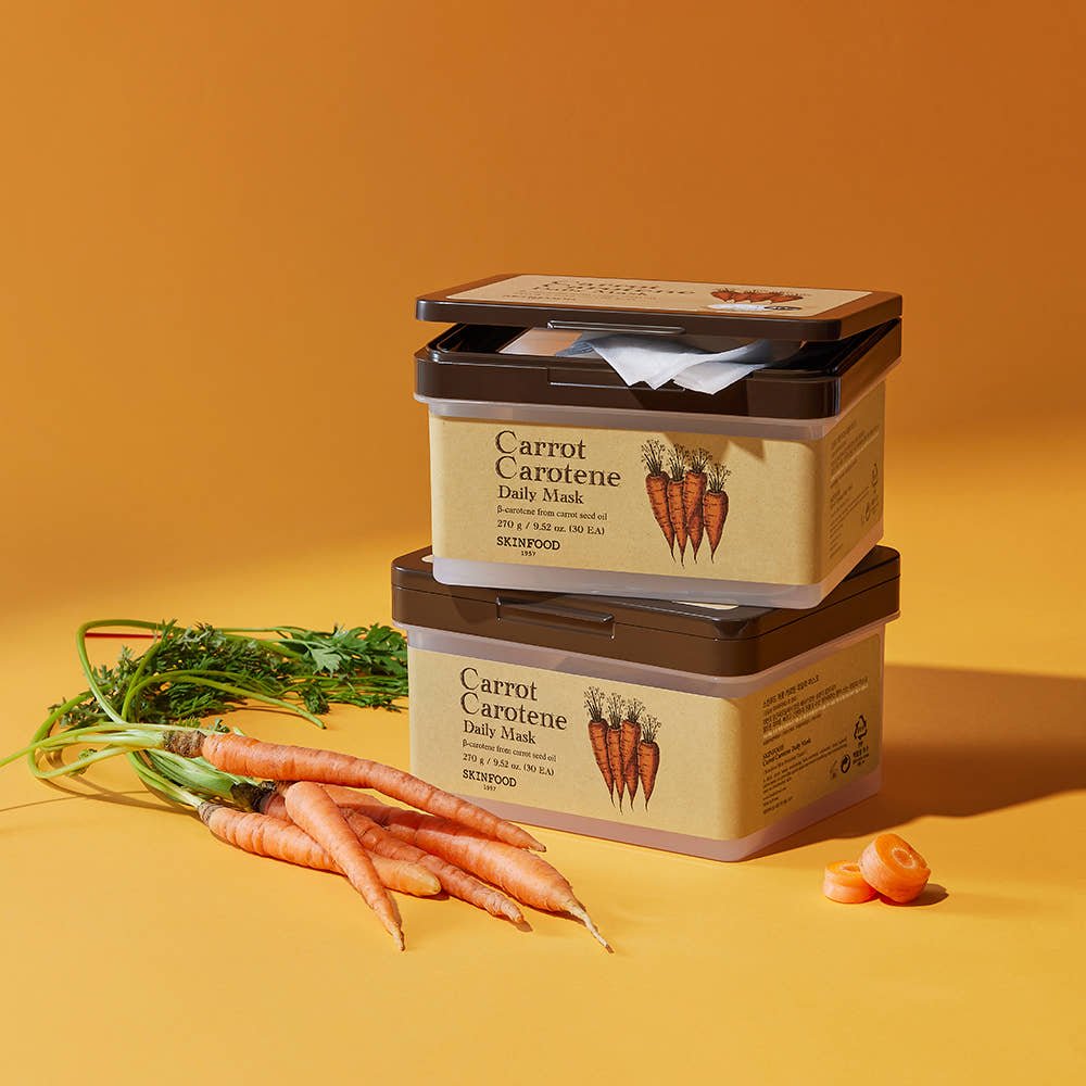 Stacked beige and brown skincare containers labeled carrot carotene daily mask with fresh carrots and carrot slices on orange background