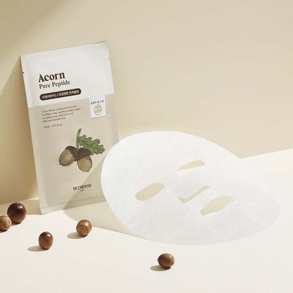 Sheet mask lying flat next to beige acorn-themed pouch with small acorns scattered on light background