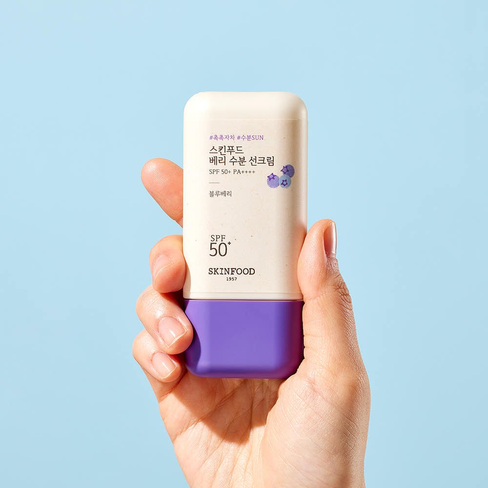 Hand holding a compact bottle of moisturizing sun cream with purple cap and berry illustration on light blue background