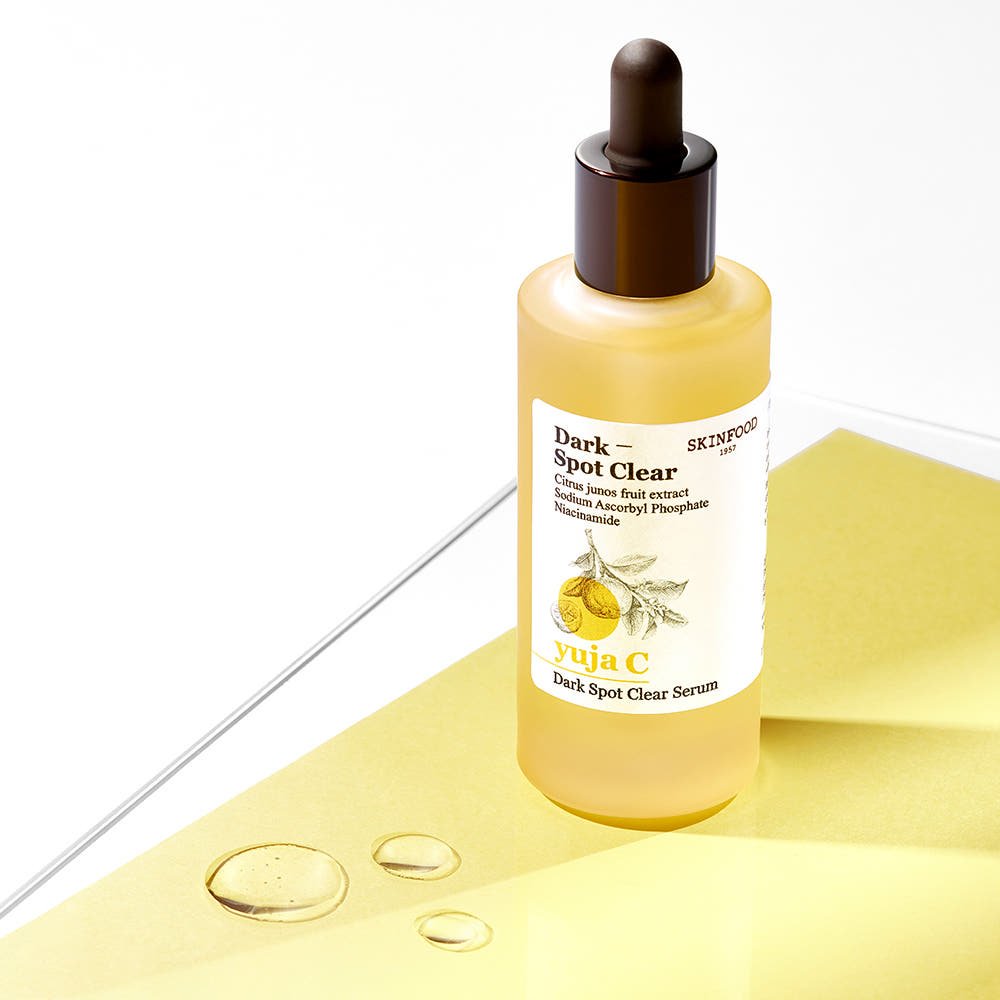 Frosted glass dropper bottle of brightening yuja C dark spot serum on yellow paper with serum droplets nearby