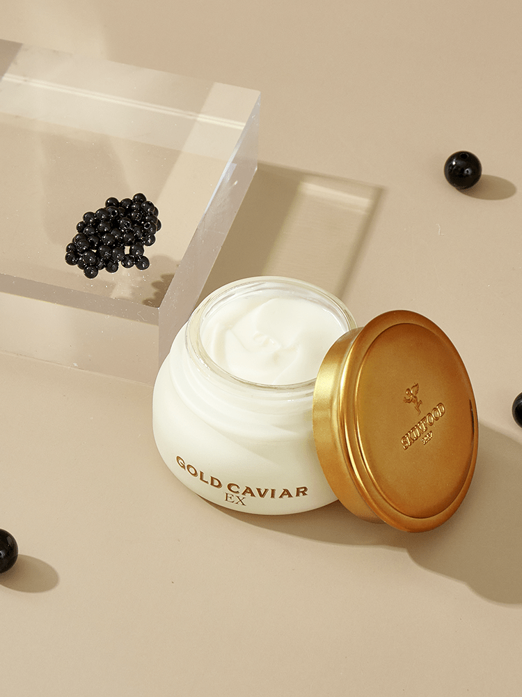 Open jar of radiant complexion cream with golden lid next to small black caviar beads on beige surface