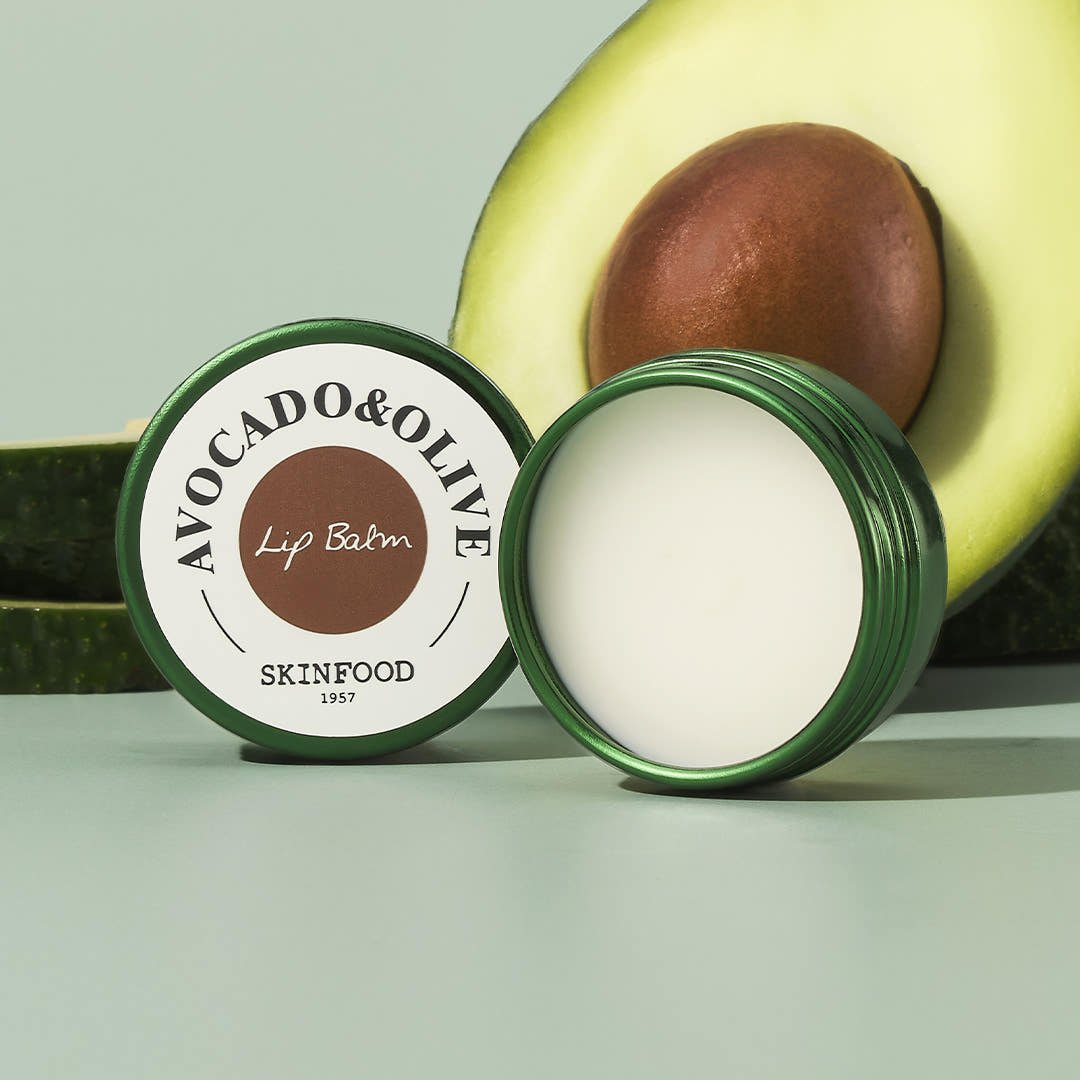 Open green jar of moisturizing avocado and olive lip balm next to halved fresh avocado on pale background