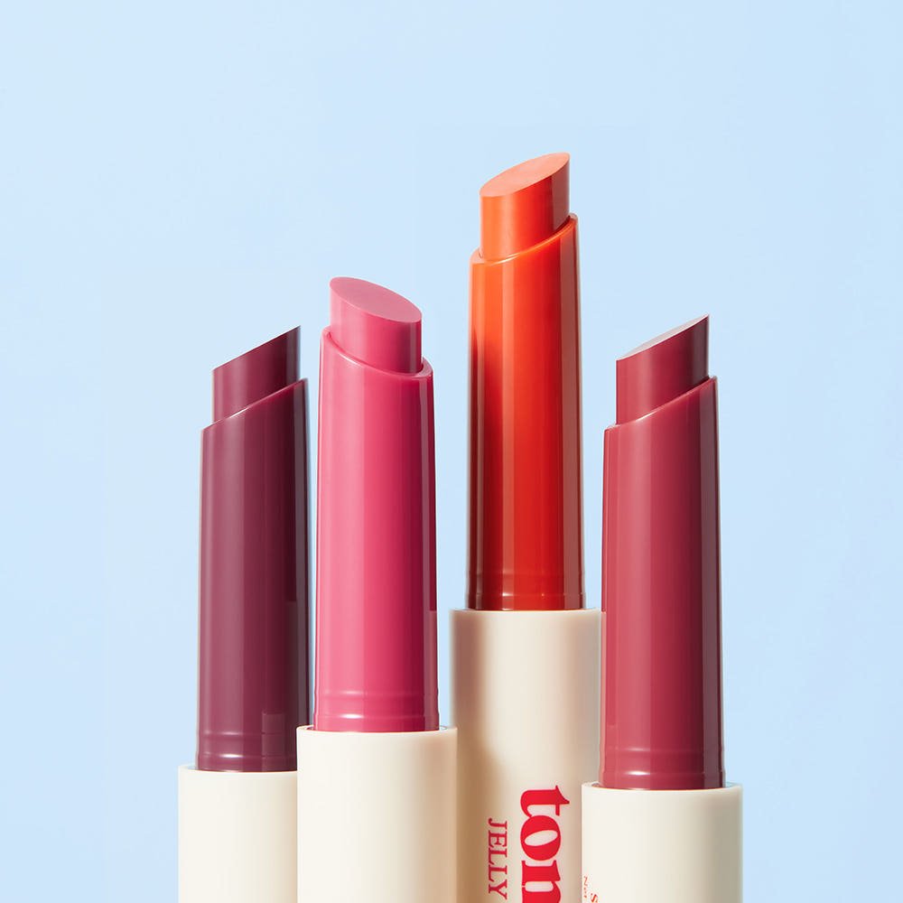 Four shades of SKINFOOD Tomato Jelly Tint Lip 1.7g in sleek white tubes showcasing vibrant colors on a soft blue background.