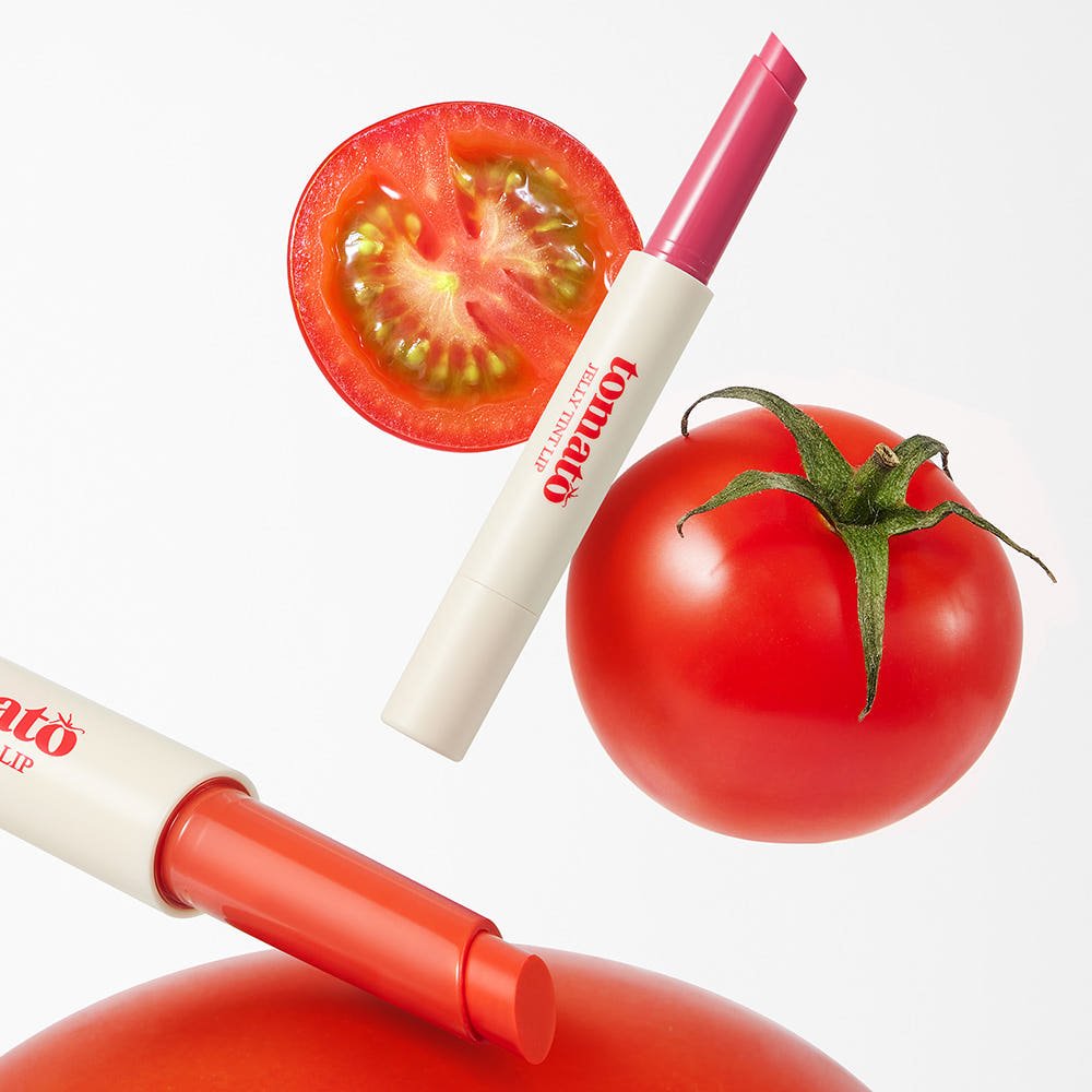 Two shades of tomato-inspired lip tint sticks next to a whole and sliced fresh tomato on white background