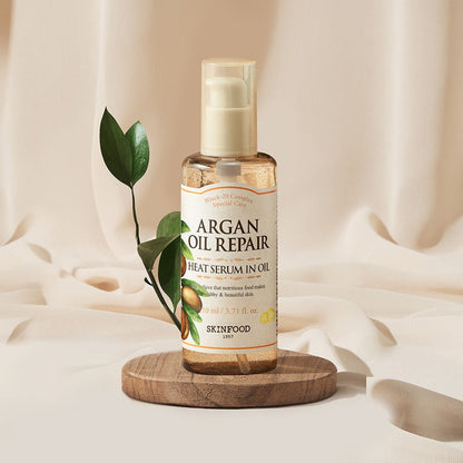 SKINFOOD Argan Oil Repair Plus Heat Serum in Oil – 110ml pump bottle on wooden platform with green leaf.