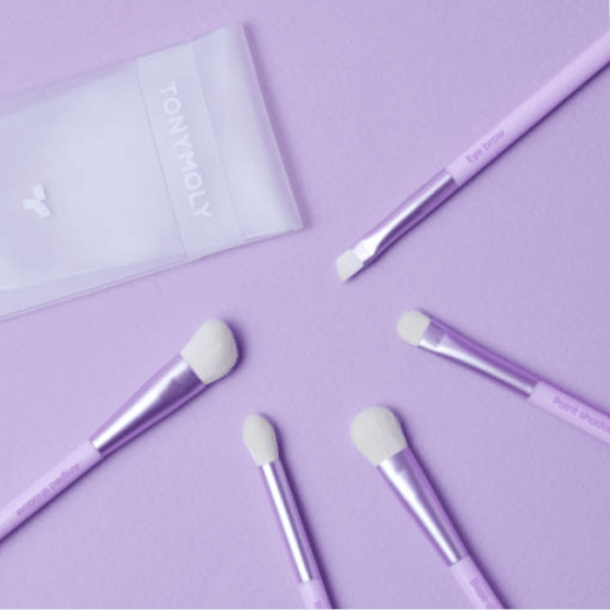 Set of five pastel purple brushes with soft white bristles arranged on matching purple surface with translucent pouch nearby