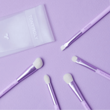 Set of five pastel purple brushes with soft white bristles arranged on matching purple surface with translucent pouch nearby