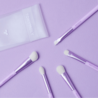 Set of five pastel purple eye makeup brushes with soft white bristles arranged on matching background with clear pouch nearby