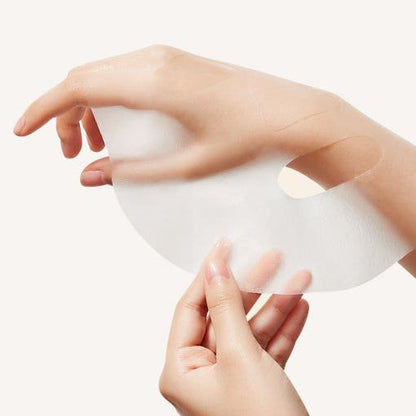 Hands holding and unfolding a translucent white jelly mask sheet against a soft beige background
