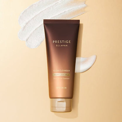 Brown gradient tube of Prestige Éclapair Glow Cleanser with creamy white cleanser swatches on beige background