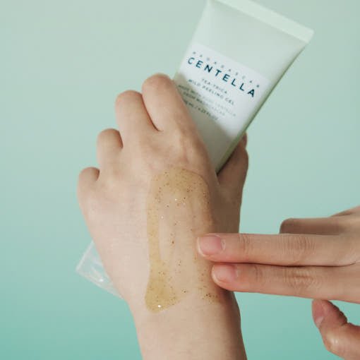 Hand applying light amber gel with visible exfoliating particles from tube on back of hand against pale blue background